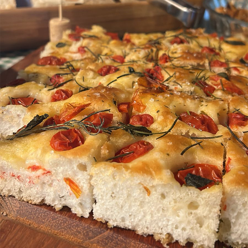 Foccacia artesanal fatiada, com casca dourada e coberta com tomates cereja assados e alecrim fresco.