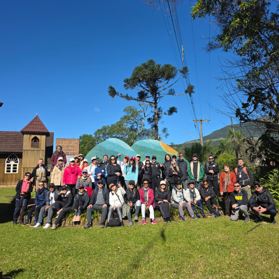Cerca de 42 caminhantes posam em Morro Azul, Três Cachoeiras, no Caminho dos Tropeiros, sob céu azul com araucárias e morros ao fundo.