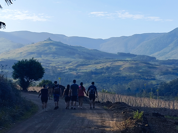 Grupo de seis trilheiros caminhando de costas por uma estrada de terra com vista para montanhas ao fundo